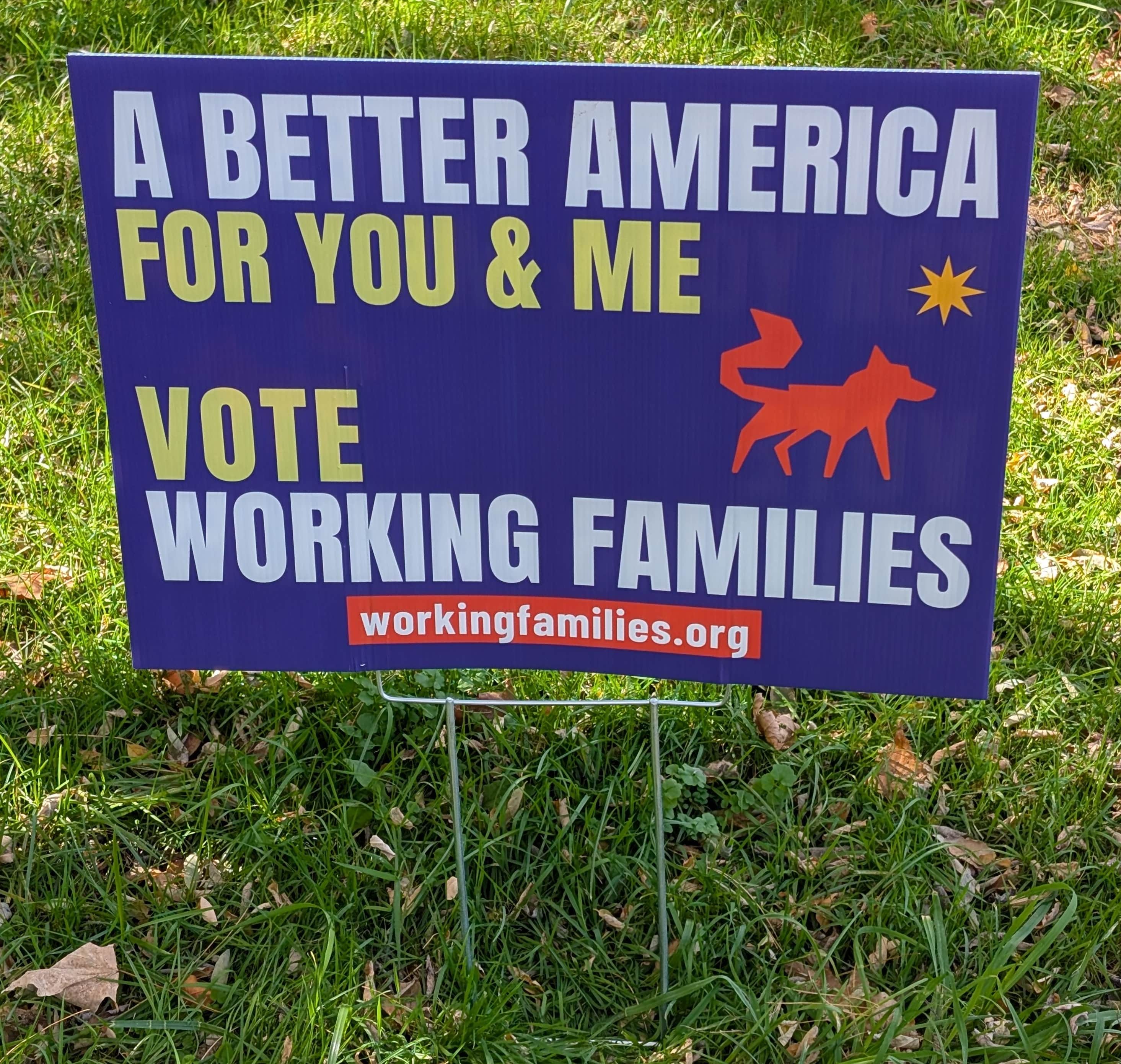 A purple yard sign staked in our lawn. It reads "A better America for you & me" and "Vote Working Families" in white and yellow, with the orange Working Families Party wolf logo, yellow north star, and workingfamilies.org on a red bar.